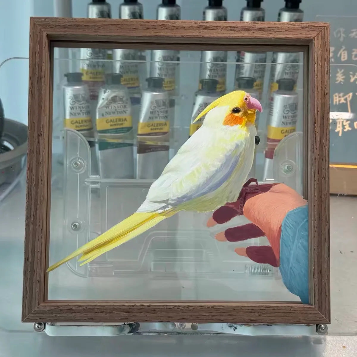 A yellow-and-white cockatiel portrait with the workshop visible through the unpainted glass.