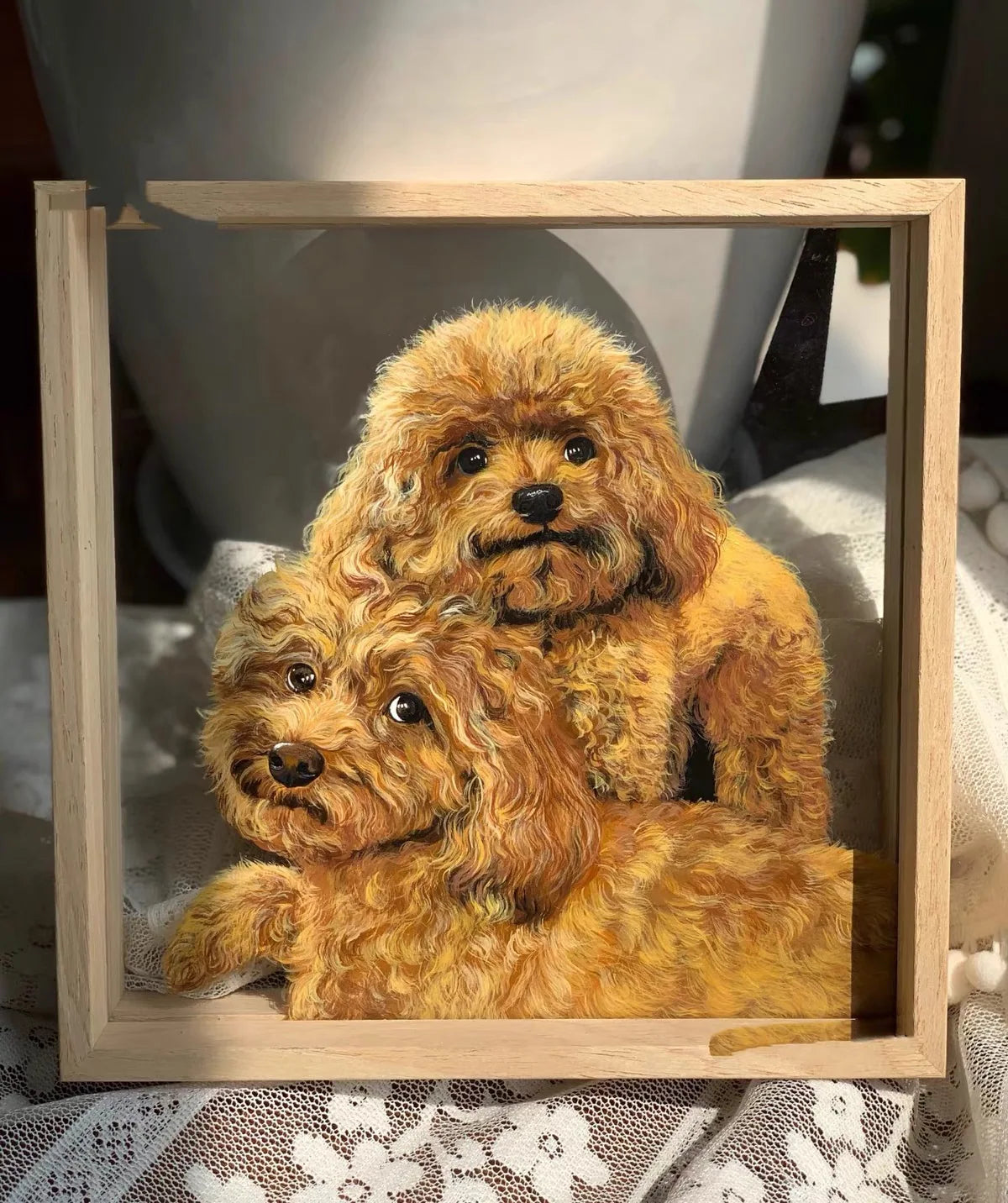 Full image shows two curly-dog portraits in one light-wood frame on a lace-covered styled tabletop, with a large white vessel/object behind. Studio setting possible; customer origin not established.