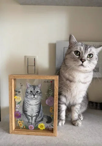 A silver tabby sits beside its floral glass portrait.